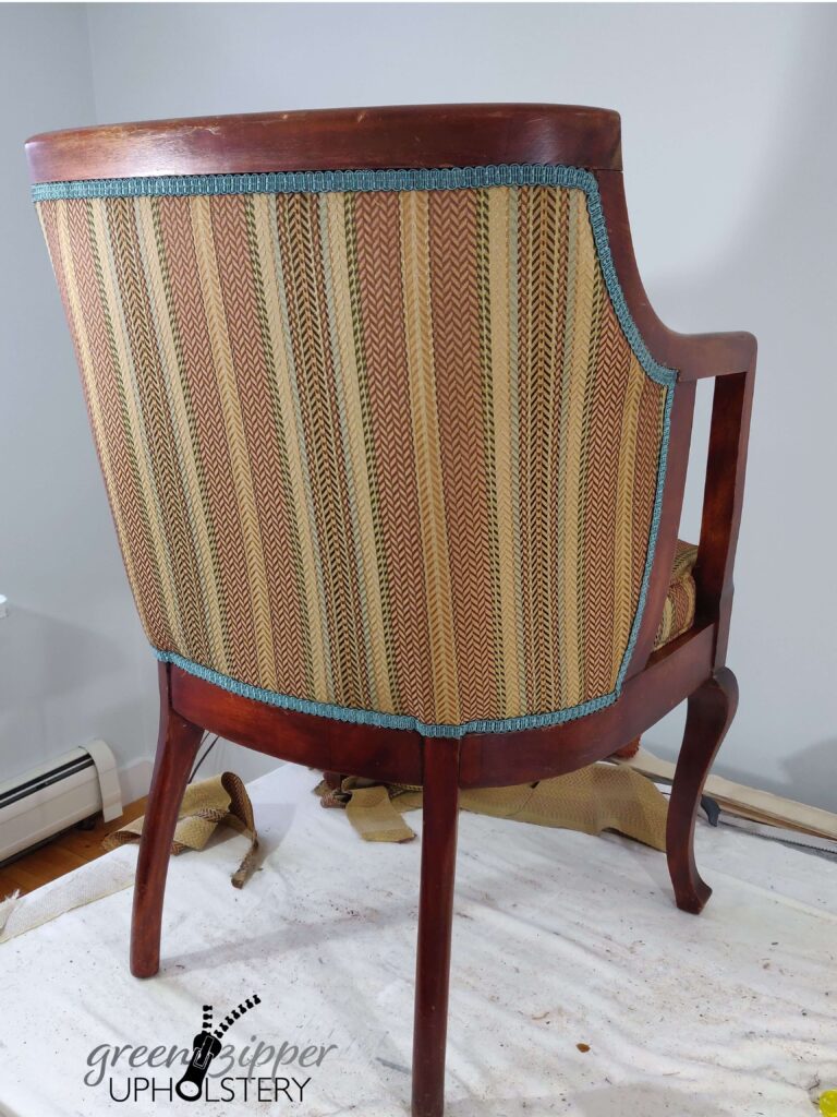 Back view of a wood frame barrel chair with a multicolor brown strip fabric with teal trim, on a work table with a green wall.