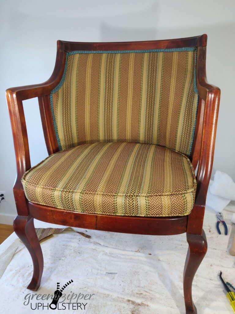 Front view of a wood frame barrel chair with a multicolor brown strip fabric with teal trim, on a work table with a green wall.