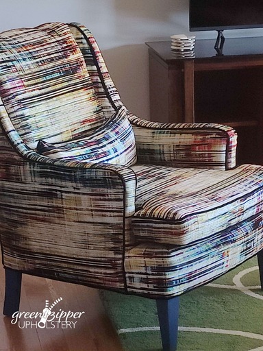 A reupholstered armchair with a graphic stripe pattern in multiple colors in living room in front of a gray wall on a wood floor with a green graphic area rug.
