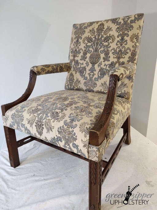 Front view of an antique chair with blue, beige and brown floral print fabric, and wood arms and legs, on a work table with a pale green wall.