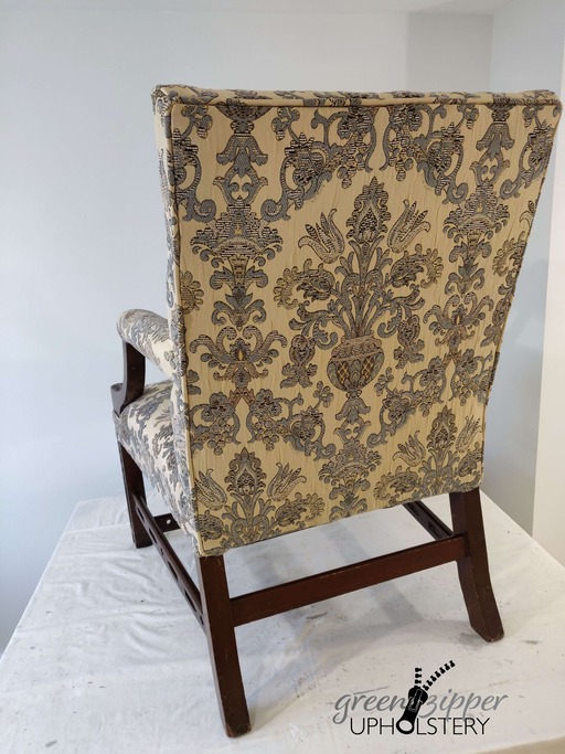 Back view of an antique chair with blue, beige and brown floral print fabric, and wood arms and legs, on a work table with a pale green wall.