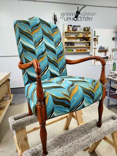Front view of a chair with wood arms and legs, and fabric in a vibrant pattern of teals, greens, browns, black and white, in a workshop.