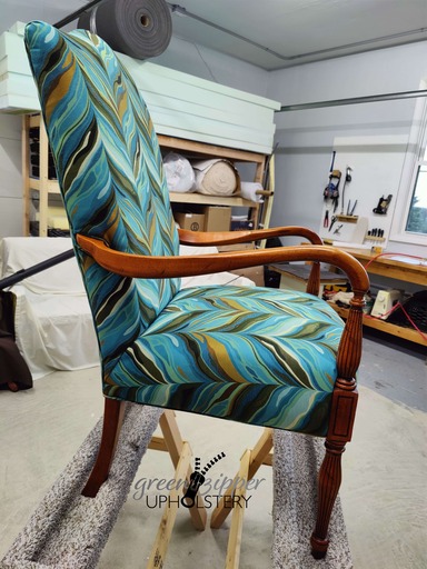 Side view of a chair with wood arms and legs, and fabric in a vibrant pattern of teals, greens, browns, black and white, in a workshop.