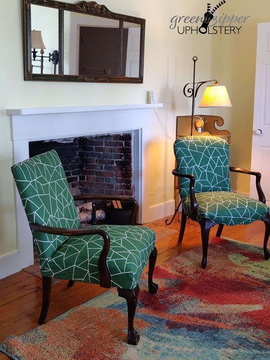 2 antique armchairs with a modern green and white fabric, in a room with yellow walls and white trim, a lamp, a yellow stand, a large antique wall mirror, and a colorful modern multicolor area rug.