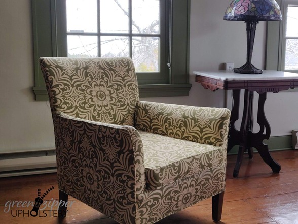 A large armchair with green and yellow floral fabric, in a room with white walls and green trim, 2 windows, a marble topped antique table with a lamp, and a wood floor.